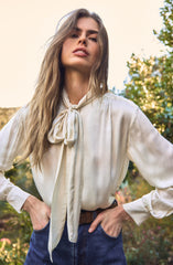A woman is wearing a cream-colored, satin blouse with long sleeves and a large bow tied at the collar. The blouse has a loose fit with pleats at the shoulders. She is also wearing high-waisted blue jeans with a brown belt.