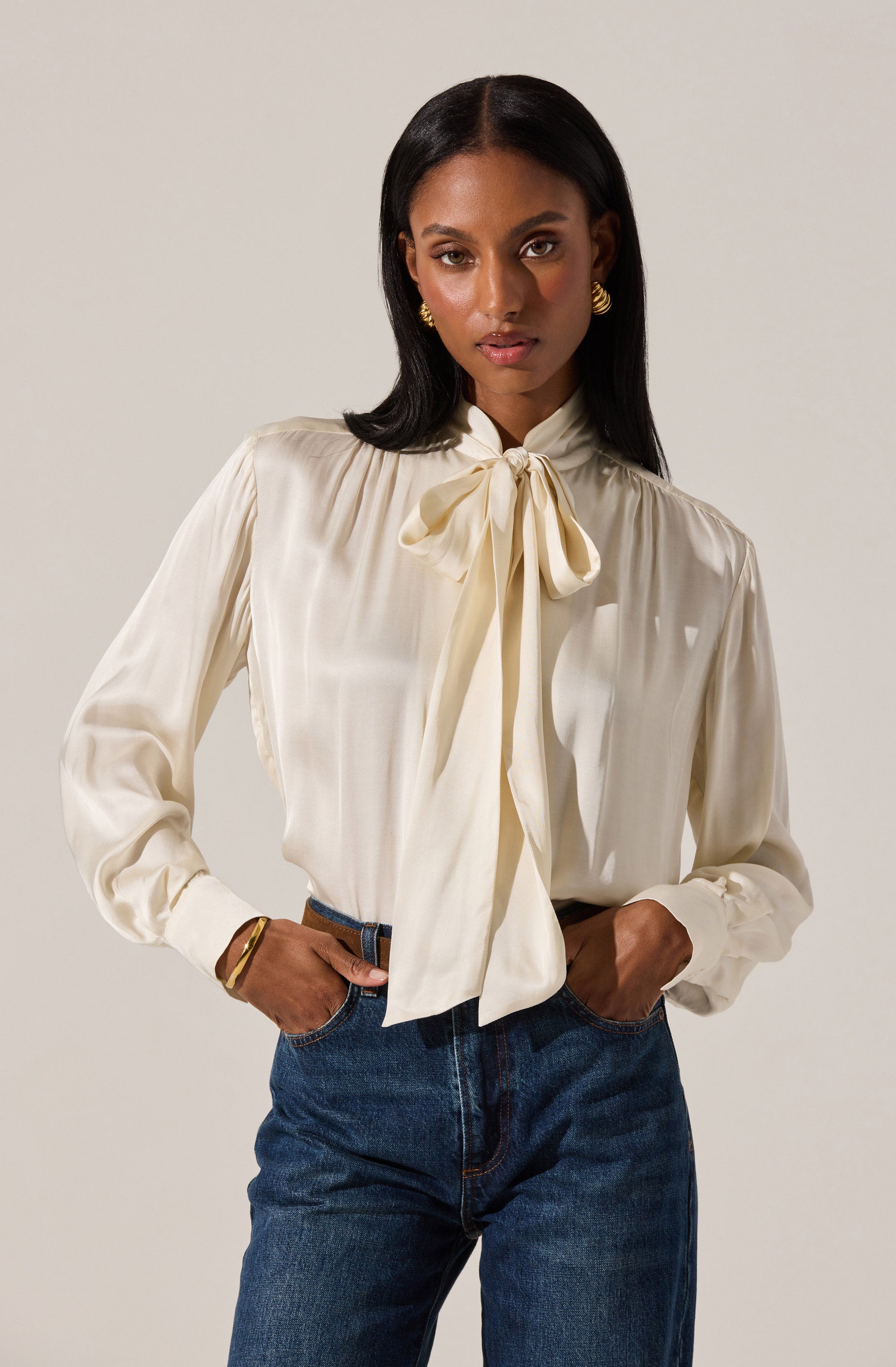 A woman is wearing a long-sleeved, cream-colored satin blouse with a large bow tied at the collar. The blouse has a slightly shiny texture, pleats around the shoulders, and puffed sleeves that gather at the cuffs. She pairs it with high-waisted, dark wash jeans. She accessorizes with gold earrings, a gold bangle on her right wrist, and a simple ring on her left hand.