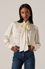 A woman is wearing a long-sleeved, cream-colored satin blouse with a large bow tied at the collar. The blouse has a slightly shiny texture, pleats around the shoulders, and puffed sleeves that gather at the cuffs. She pairs it with high-waisted, dark wash jeans. She accessorizes with gold earrings, a gold bangle on her right wrist, and a simple ring on her left hand.