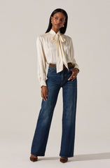 A woman is wearing a cream-colored blouse with long sleeves that have buttoned cuffs. The blouse features a large bow tied at the collar. She pairs the blouse with high-waisted, wide-leg blue jeans and brown shoes. She accessorizes with gold hoop earrings, a gold bracelet, and a ring.