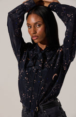 A woman is wearing a long-sleeved, dark satin blouse with a whimsical celestial print featuring stars, clouds, and crescent moons in light-colored shades. The blouse has a large bow tied at the collar. She pairs it with black jeans and a matching black belt.