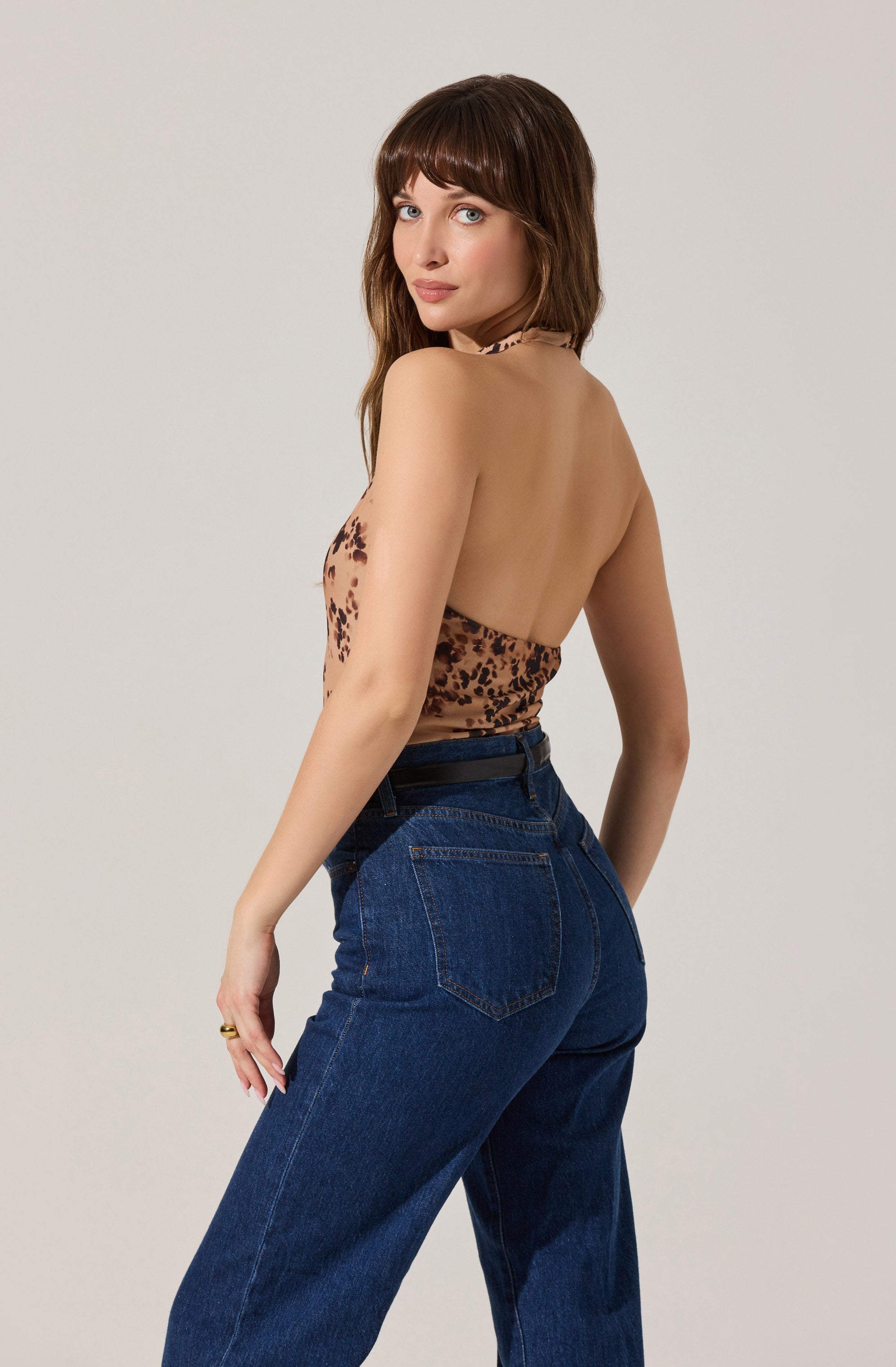 A woman with shoulder-length brown hair and bangs is wearing a halter top with a brown and black leopard print pattern. She is also wearing high-waisted, dark blue denim jeans. She is standing against a plain light gray background, turned slightly to the side with her head turned toward the camera.