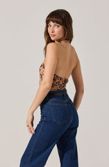 A woman with shoulder-length brown hair and bangs is wearing a halter top with a brown and black leopard print pattern. She is also wearing high-waisted, dark blue denim jeans. She is standing against a plain light gray background, turned slightly to the side with her head turned toward the camera.