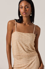 A woman is wearing a sleeveless, beige dress with thin spaghetti straps. The dress has a textured look with small, shiny embellishments all over the fabric.