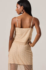 A woman is wearing a sleeveless, beige, sequined dress with thin spaghetti straps. The dress has a fitted top and a sheer, sequined skirt that extends below the knee. She is accessorized with large pearl drop earrings.