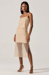 The woman is wearing a sleeveless, beige dress with thin spaghetti straps. The top part of the dress is fitted and made of a sparkling, textured fabric. The skirt portion is high-waisted and short, ending above the knees, with an outer layer of sheer, sparkly fabric that extends to mid-calf length and features a thigh-high slit. She is also wearing beige high-heeled sandals with ankle straps and pearl drop earrings.