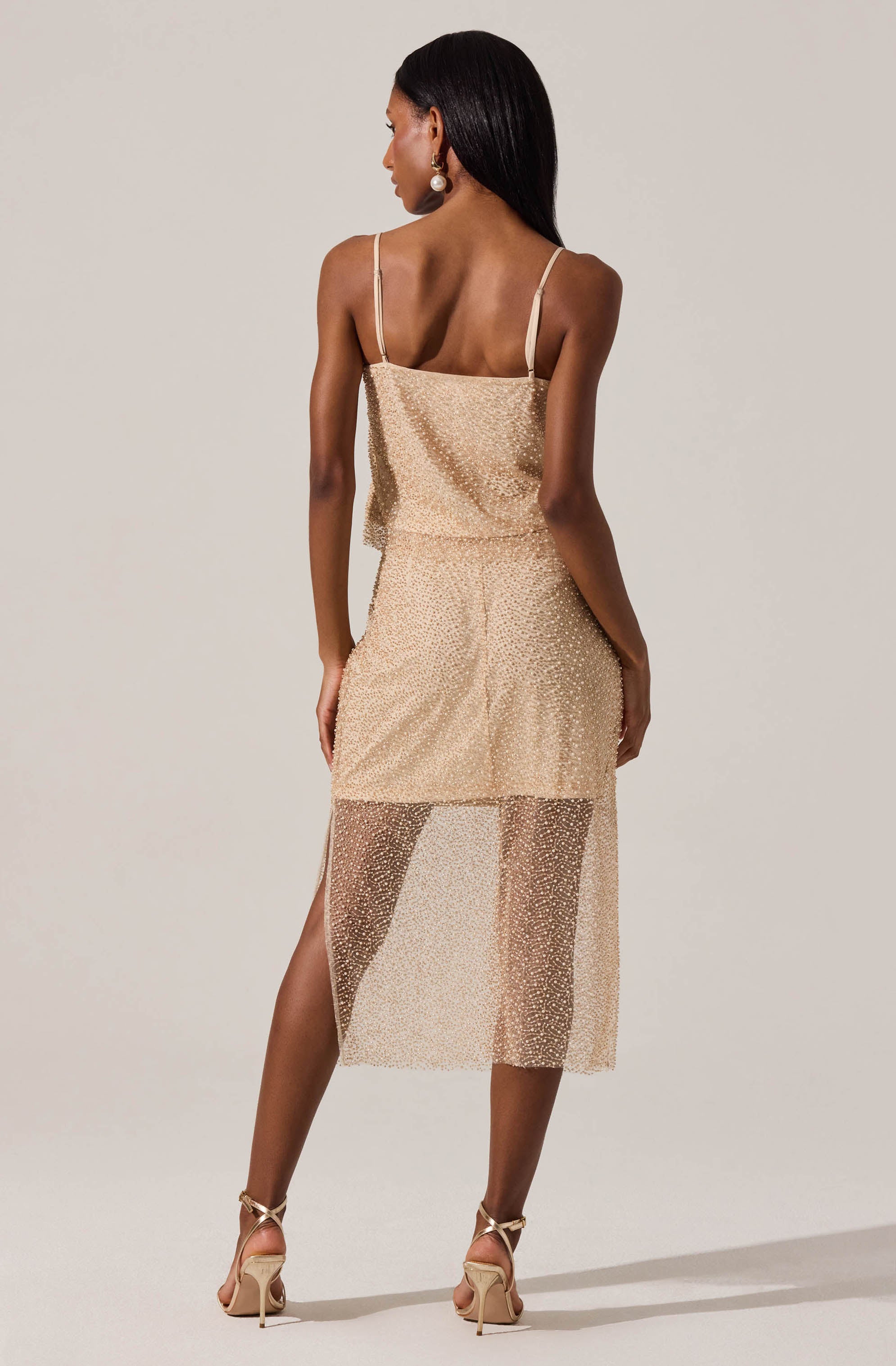 A woman is wearing a two-piece dress with a fitted, glittering gold top with thin spaghetti straps and a matching gold skirt. The skirt is knee-length with an overlay of sheer, glittery fabric that extends below the solid layer, creating a semi-transparent look. The woman is also wearing high-heeled sandals and large pearl earrings.