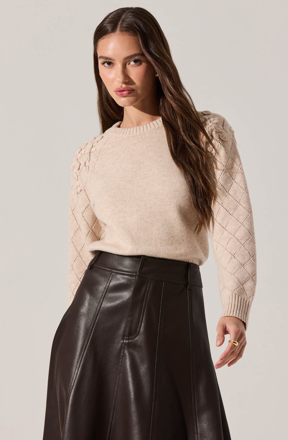 A woman is wearing a light beige knit sweater with diamond-shaped textured patterns on the sleeves and shoulders. She pairs it with a high-waisted, dark brown leather skirt that features visible stitching details.
