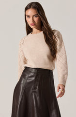 A woman is wearing a light beige knit sweater with diamond-shaped textured patterns on the sleeves and shoulders. She pairs it with a high-waisted, dark brown leather skirt that features visible stitching details.