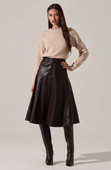 A woman is wearing a beige knit sweater with textured diamond patterns on the shoulders and sleeves. She pairs it with a high-waisted, dark brown leather A-line skirt that falls below the knees. She completes the outfit with tall, black leather thigh-high boots.