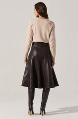 A woman is wearing a light beige sweater with textured diamond patterns on the sleeves and shoulders. She pairs it with a high-waisted, dark brown leather skirt that flares out below the waist. She completes the outfit with tall, matching dark brown leather thigh-high boots with high heels.