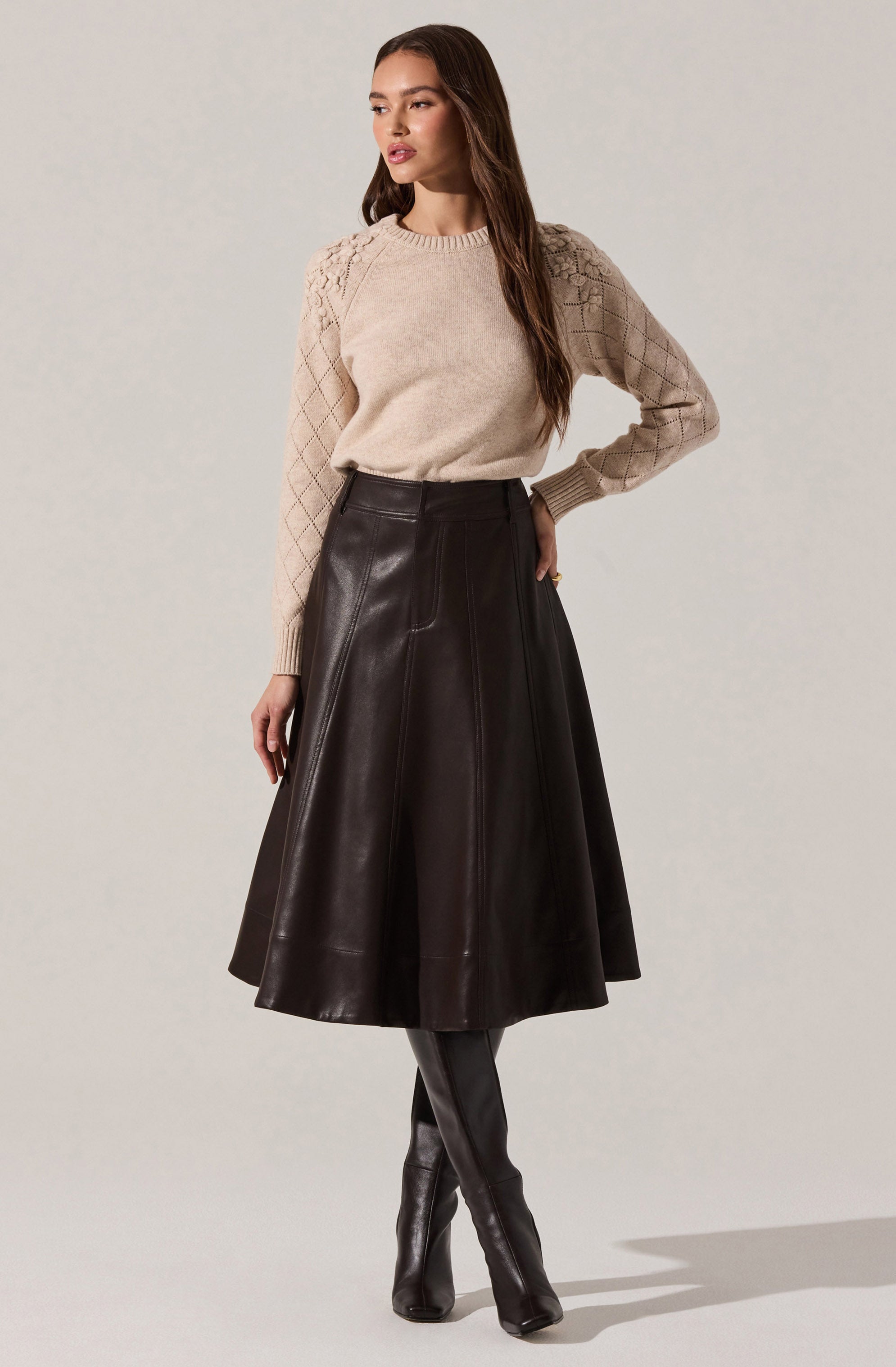 A woman is wearing a beige knit sweater with a textured pattern on the shoulders and sleeves, paired with a high-waisted, knee-length, dark brown leather skirt that has visible stitching details. She completes the look with black knee-high leather boots.
