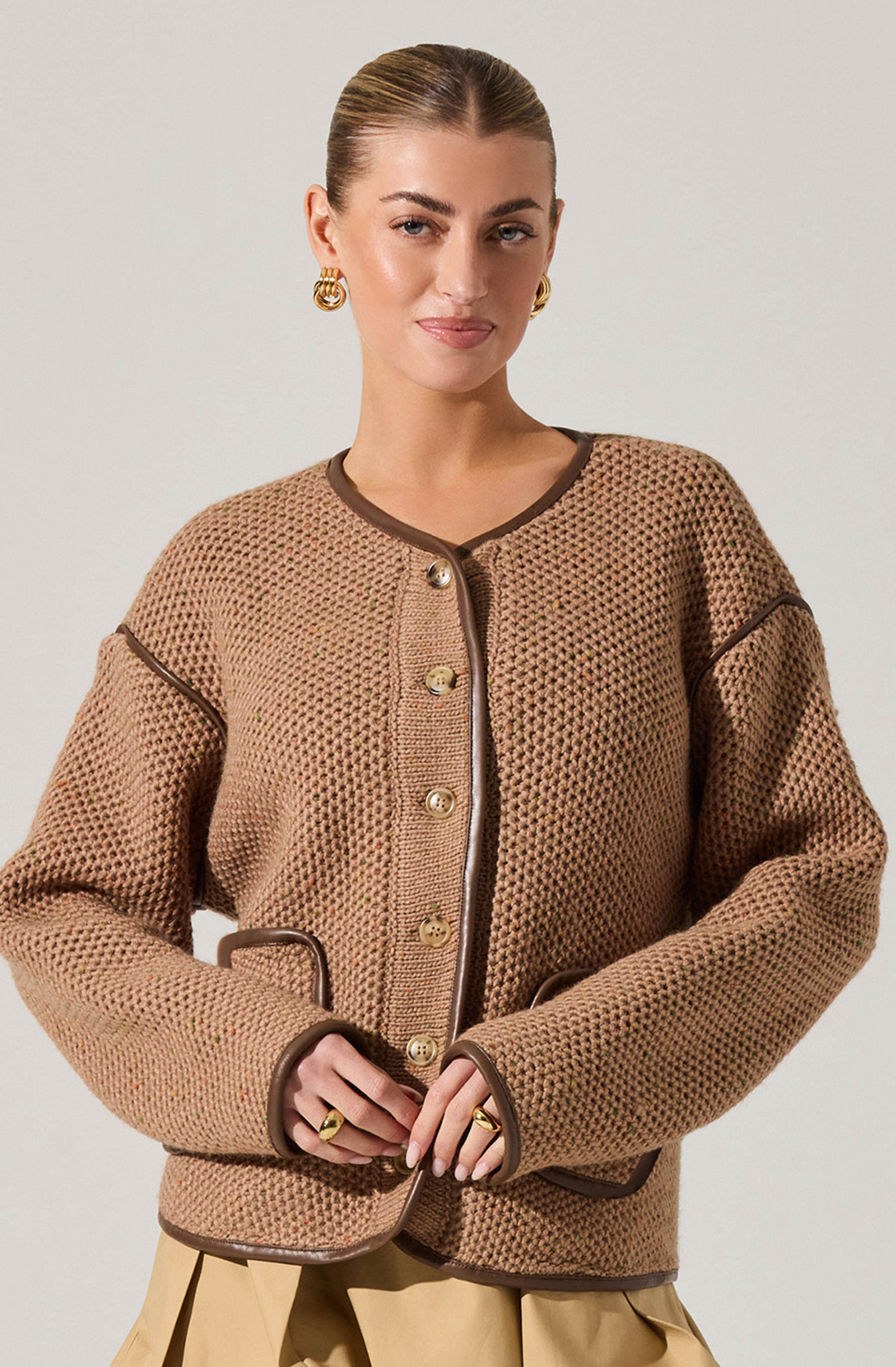 A woman is wearing a beige, textured knit jacket with dark brown trim along the collar, front opening, pockets, and sleeve seams. The jacket features gold buttons down the front and pockets, with the trim adding contrast.