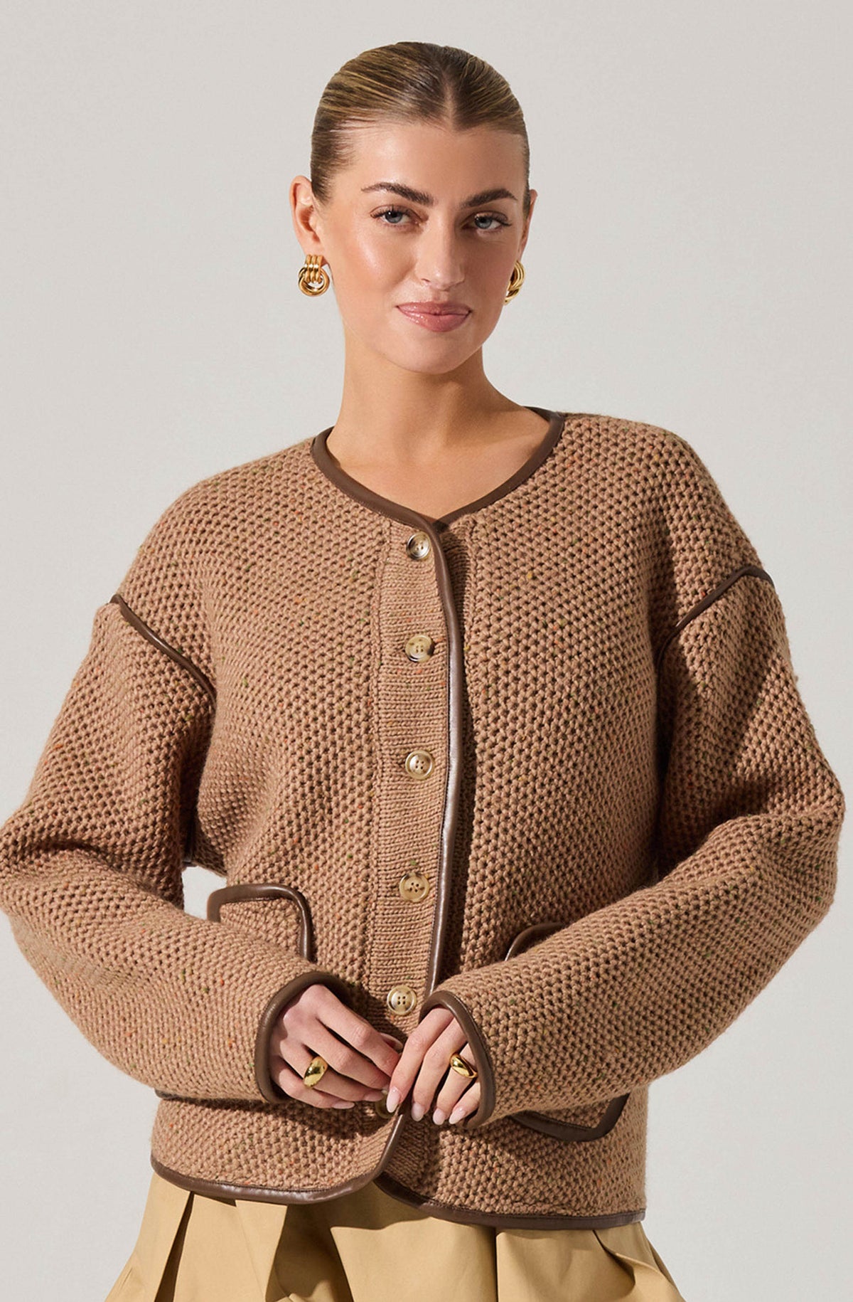 A woman is wearing a beige, textured knit jacket with dark brown trim along the collar, front opening, pockets, and sleeve seams. The jacket features gold buttons down the front and pockets, with the trim adding contrast.