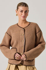 A woman is wearing a beige, textured knit jacket with dark brown trim along the collar, front opening, pockets, and sleeve seams. The jacket features gold buttons down the front and pockets, with the trim adding contrast.