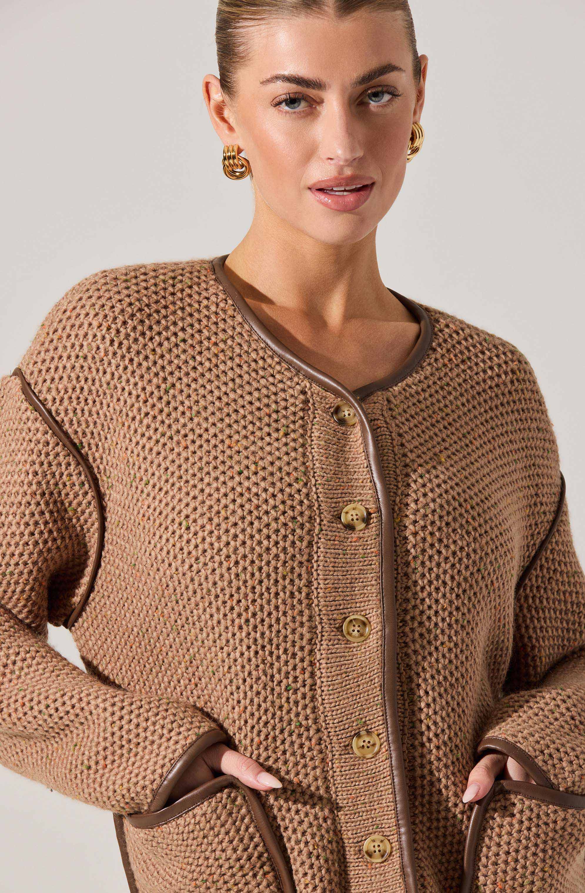 A woman is wearing a light brown, textured knit jacket with a rounded neckline. The jacket features large, round, shiny gold buttons down the front and metallic trim along the edges of the collar, pockets, and sleeve cuffs. She is accessorizing with large, gold hoop earrings.