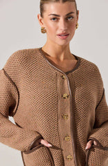 A woman is wearing a light brown, textured knit jacket with a rounded neckline. The jacket features large, round, shiny gold buttons down the front and metallic trim along the edges of the collar, pockets, and sleeve cuffs. She is accessorizing with large, gold hoop earrings.
