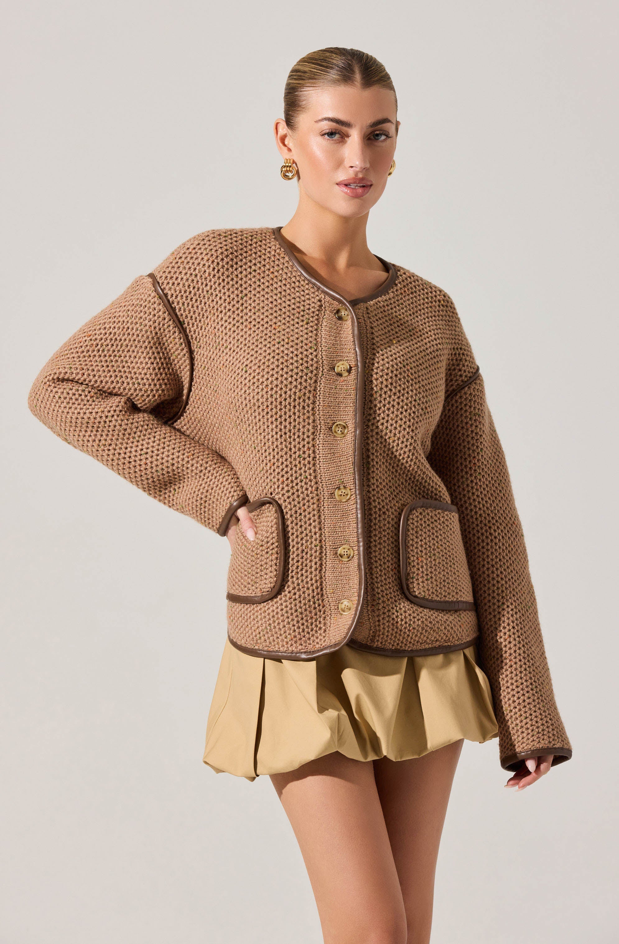 A woman is wearing a tan, textured knit cardigan with dark brown piping along the edges and pockets, and gold buttons down the front. She is also wearing beige shorts with a loose, gathered design.