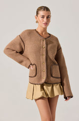 A woman is wearing a tan, textured knit cardigan with dark brown piping along the edges and pockets, and gold buttons down the front. She is also wearing beige shorts with a loose, gathered design.
