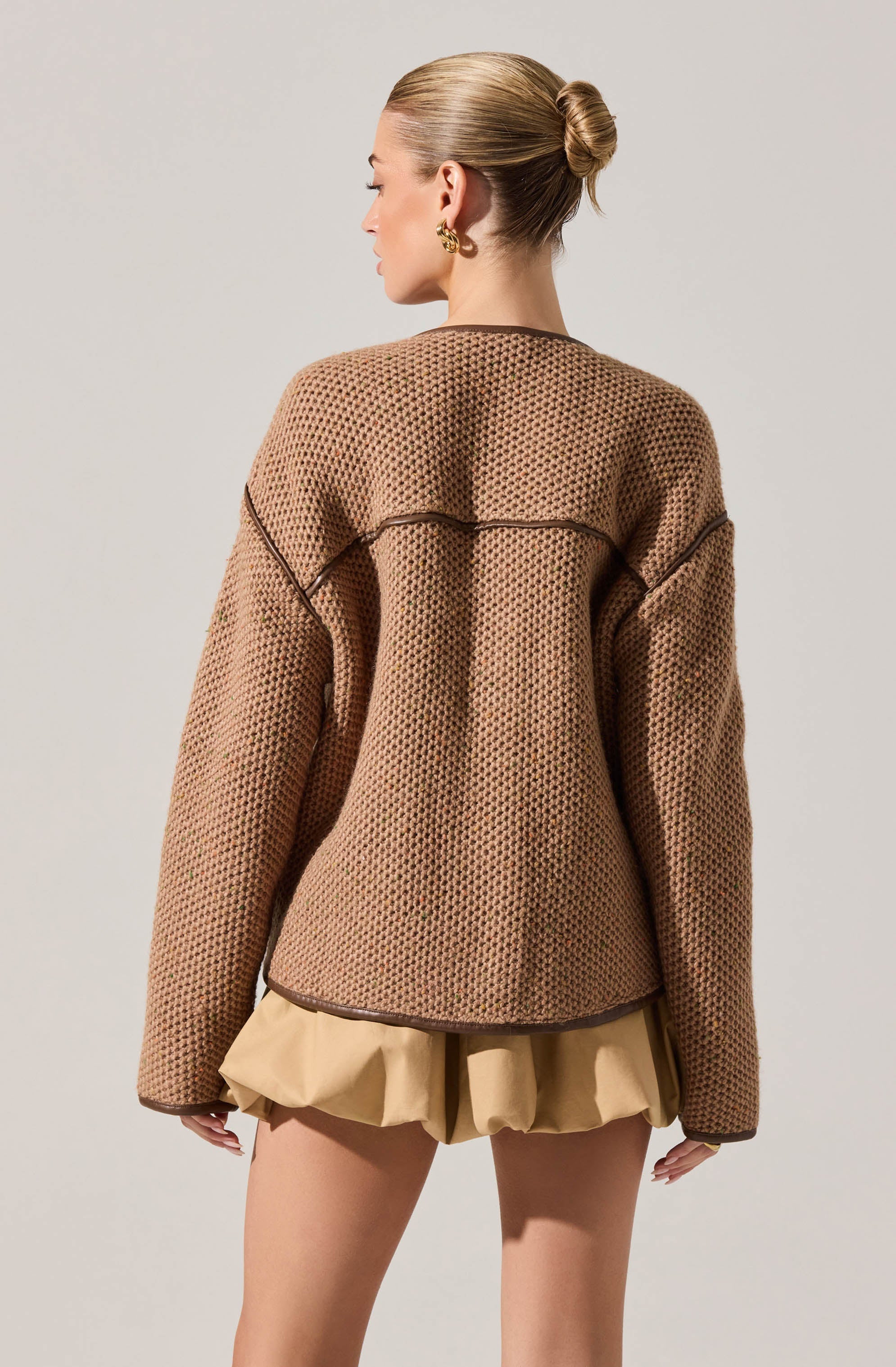 A woman is wearing a loose, oversized, brown textured jacket with a honeycomb pattern and dark trim seams. She pairs it with beige, gathered shorts.