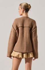 A woman is wearing a loose, oversized, brown textured jacket with a honeycomb pattern and dark trim seams. She pairs it with beige, gathered shorts.