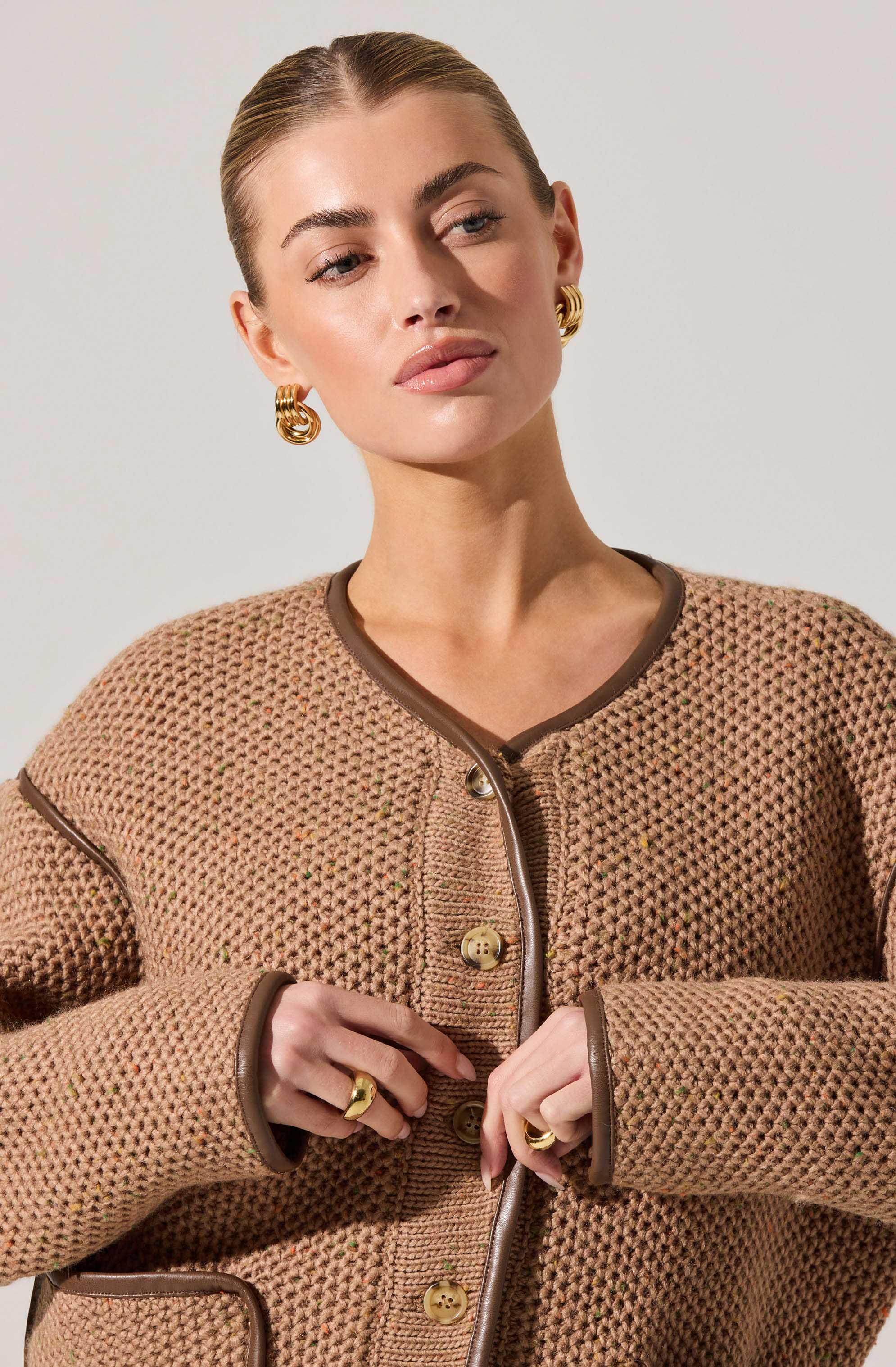 A woman wearing a light brown, textured, knit jacket with dark brown piping trim along the edges and cuffs. The jacket features large, decorative buttons down the front. She is accessorized with gold hoop earrings and multiple rings.