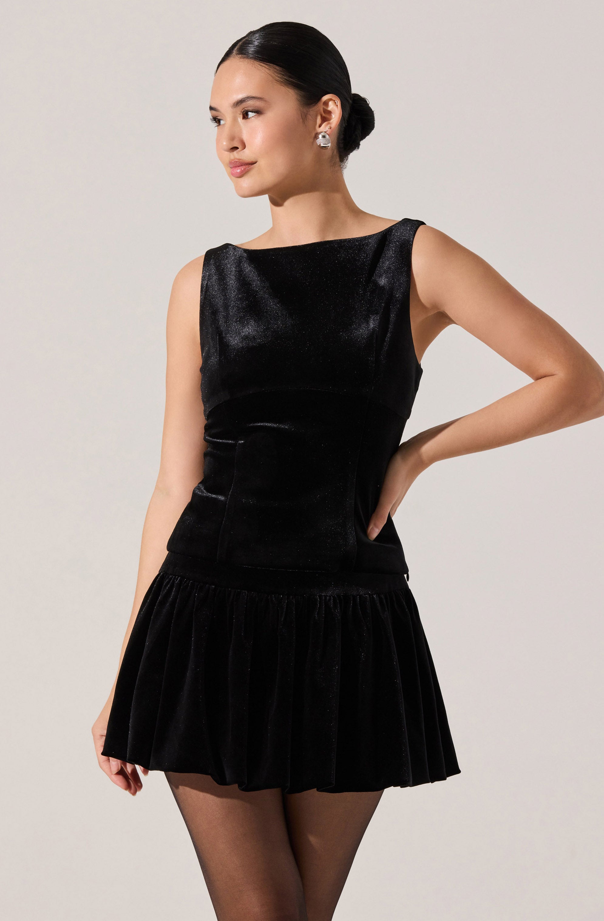 A woman is wearing a sleeveless black velvet dress with a fitted bodice and a rounded neckline. The dress has a short, pleated skirt that adds volume and texture to the outfit. The woman is accessorized with large, round earrings and has her hair styled in a sleek, low bun.