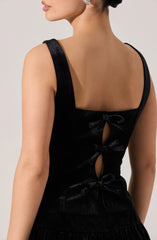 A woman is wearing a sleeveless black dress with wide shoulder straps. The back of the dress features three vertical cutouts, each tied with a small black bow. The fabric of the dress has a subtle shiny texture.