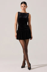 A woman is wearing a sleeveless black dress made of a shiny, velvety fabric with a fitted bodice and a gathered, ruffled skirt that falls above the knees. She has paired the outfit with black sheer tights and black high heels adorned with white polka dots.