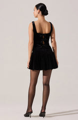 A woman is wearing a sleeveless black dress with wide shoulder straps, a fitted bodice with cut-out details in the back that are tied with small bows, and a short, flared skirt made of a sheer, lightweight fabric. She is also wearing black sheer stockings and black high-heeled shoes decorated with small silver studs.