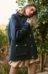 A woman is wearing a navy blue jacket with large gold buttons and a brown collar. The jacket has two front pockets with gold button flaps. She is also wearing beige, puffy shorts with a gathered, bubble-like appearance. The background shows lush green foliage and a partly cloudy sky.