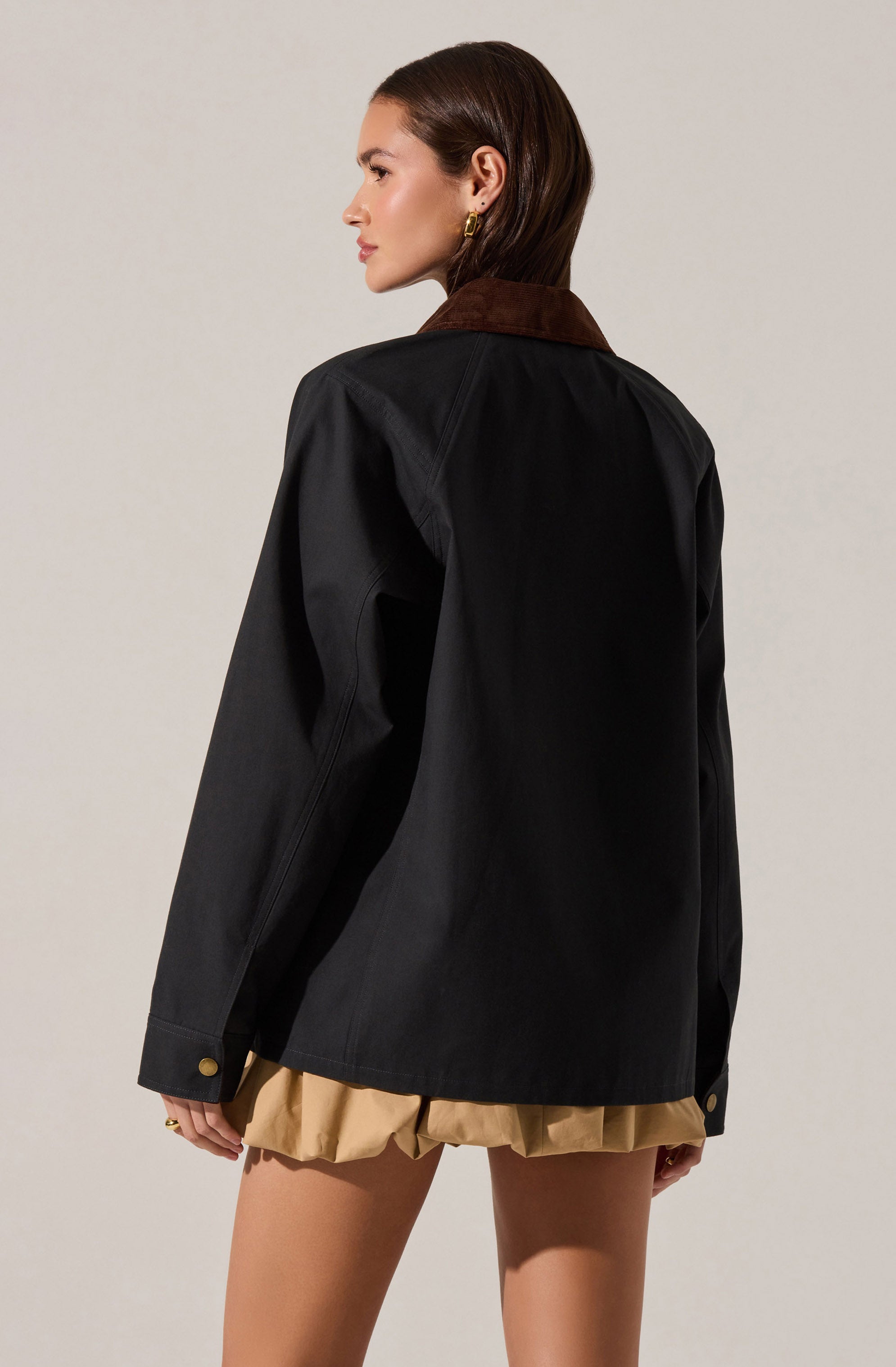 A woman wearing a black jacket with a brown collar, featuring long sleeves with buttoned cuffs, and beige shorts with a ruffled hem.