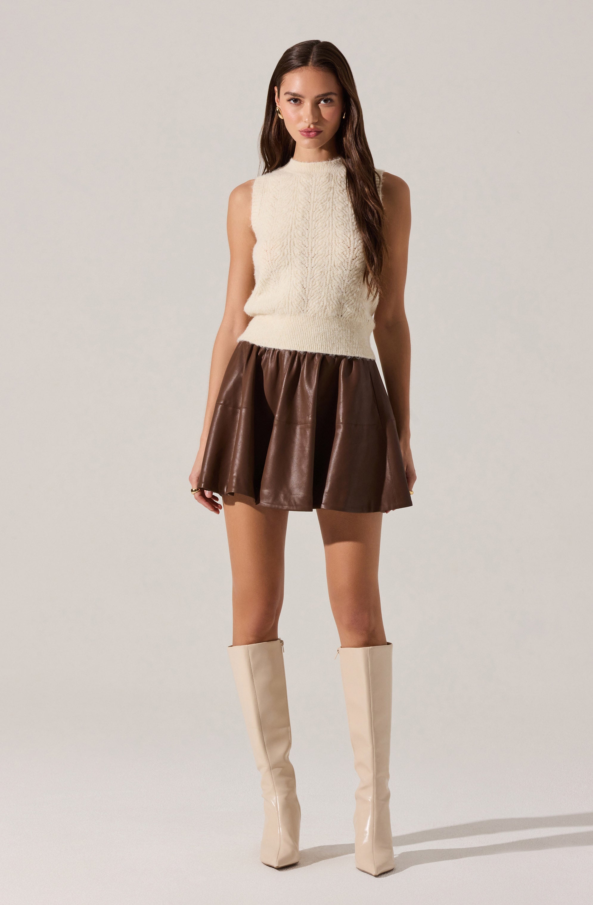 A woman is wearing a sleeveless, high-neck, cream-colored knit top and a short, voluminous brown leather skirt. She has on tall, cream-colored boots that reach just below her knees.