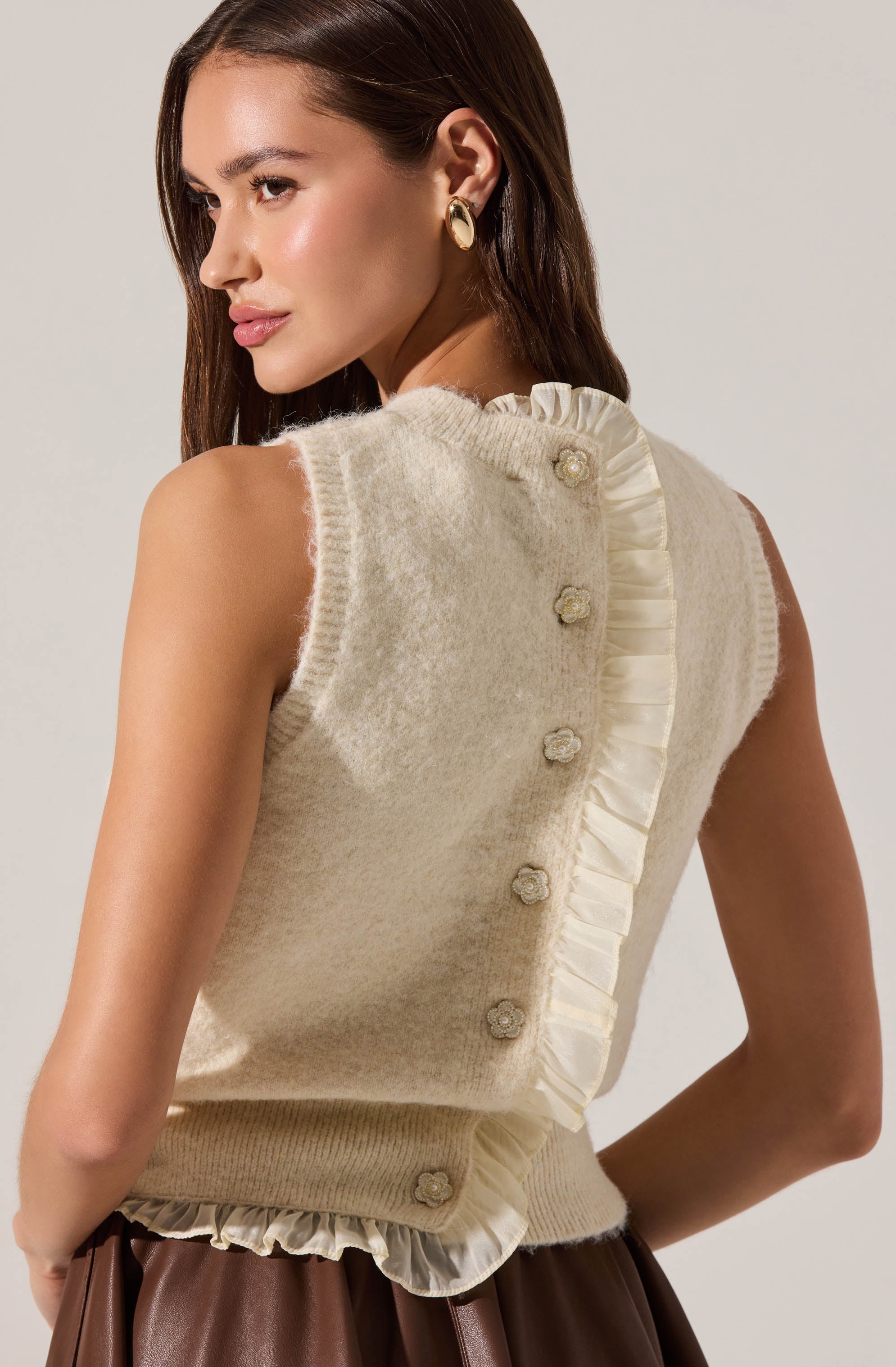 A woman is wearing a sleeveless, cream-colored knitted vest with a soft, fuzzy texture. The vest features decorative fabric ruffles along the back and neckline, with small, fabric flower buttons running down the back. She pairs it with a brown skirt or pants and accessorizes with large, gold hoop earrings.