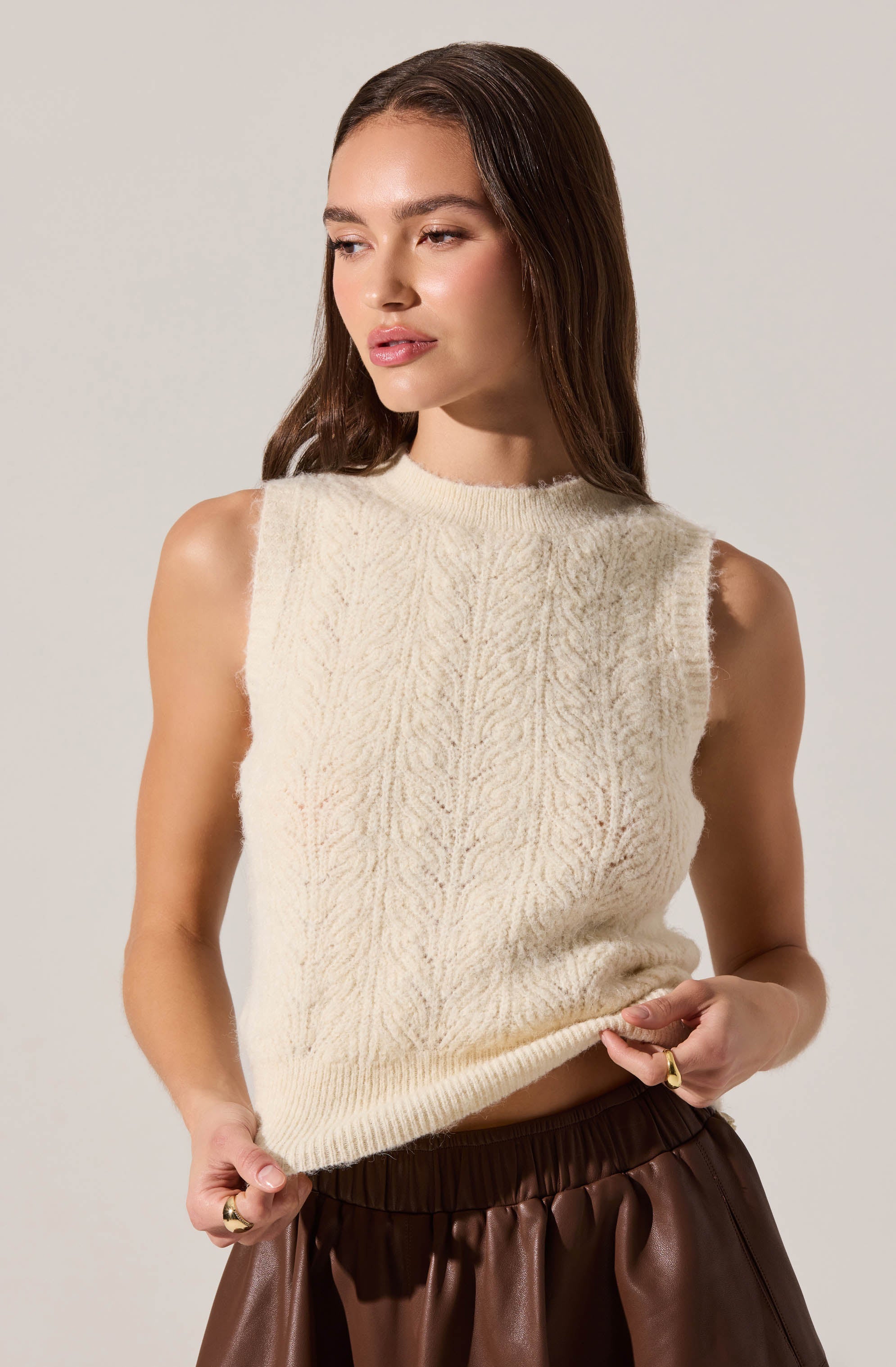 A woman is wearing a sleeveless, cream-colored knitted vest with a subtle textured pattern. The vest has a ribbed neckline and hem, and the armholes have a slightly frayed edge. She pairs it with dark brown, shiny, high-waisted pants.