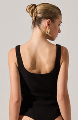 A woman is wearing a sleeveless, black bodysuit with wide straps and a square neckline. Her hair is styled in a tight bun, and she has large, gold, crescent-shaped earrings.