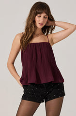A woman is wearing a sleeveless, burgundy pleated top with thin spaghetti straps. The top has a loose, flowy fit and a banded hem at the bottom. She pairs it with black shorts that have small, shiny embellishments, and sheer black tights.
