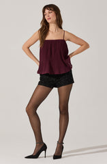 A woman is wearing a maroon, pleated, spaghetti strap top with a square neckline. She has on black shorts that feature small, scattered rhinestones or specks. She is also wearing sheer black tights and black high-heeled shoes with ankle straps.