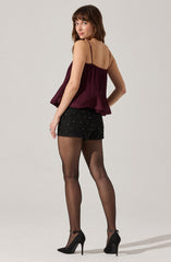 A woman is wearing a sleeveless, burgundy pleated top with thin spaghetti straps. She pairs the top with black shorts decorated with small, shiny embellishments. She completes the outfit with sheer black tights and black high-heeled shoes with ankle straps.