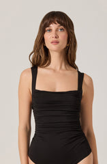 A woman is wearing a black sleeveless bodysuit with wide shoulder straps and a gathered, ruched detail across the front.