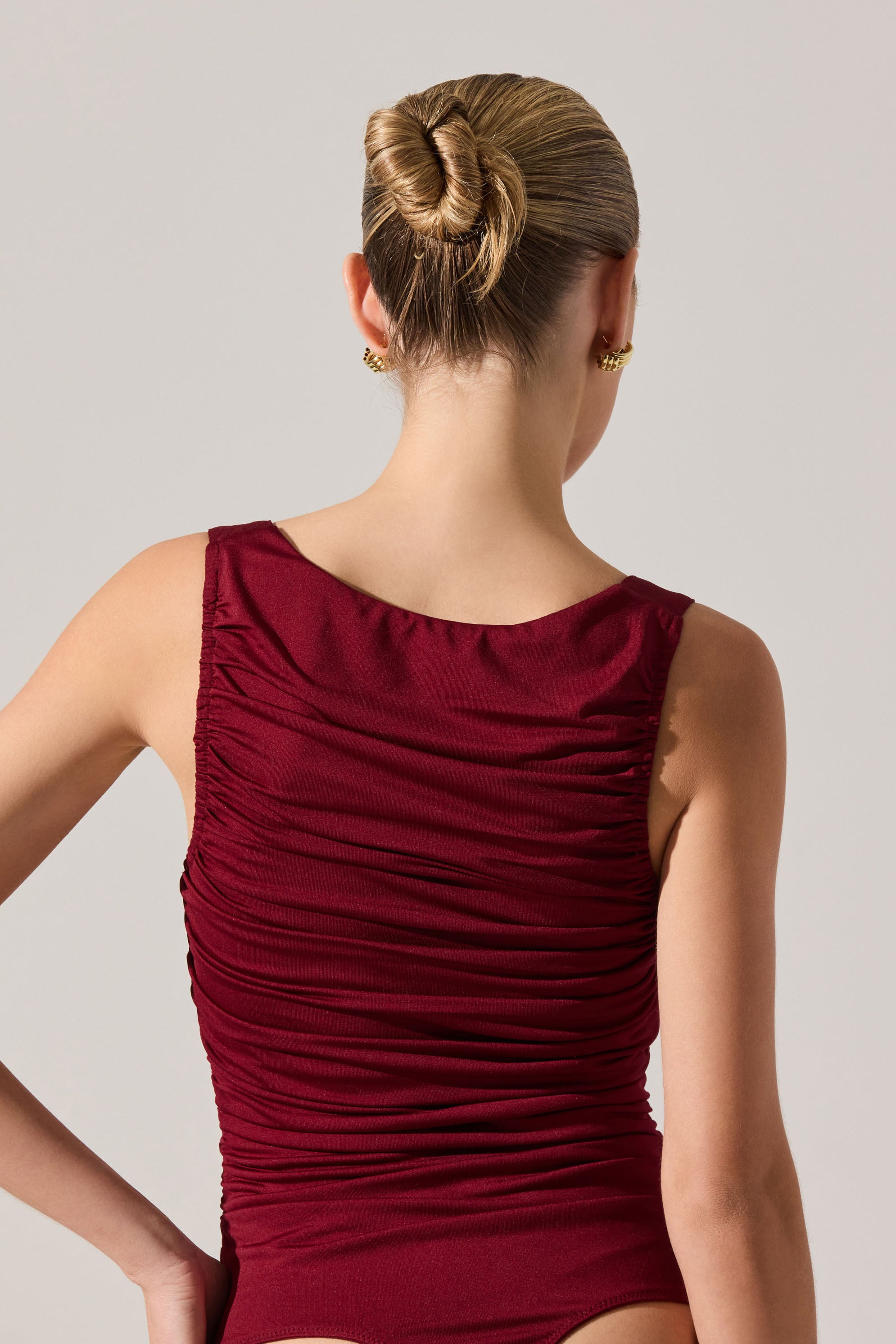 A woman is wearing a sleeveless, deep red or burgundy dress with a rounded neckline. The dress features a gathered, ruched fabric design that creates texture and visual interest across the front and back. She has her hair styled in a smooth, tight bun and is accessorized with gold stud earrings.