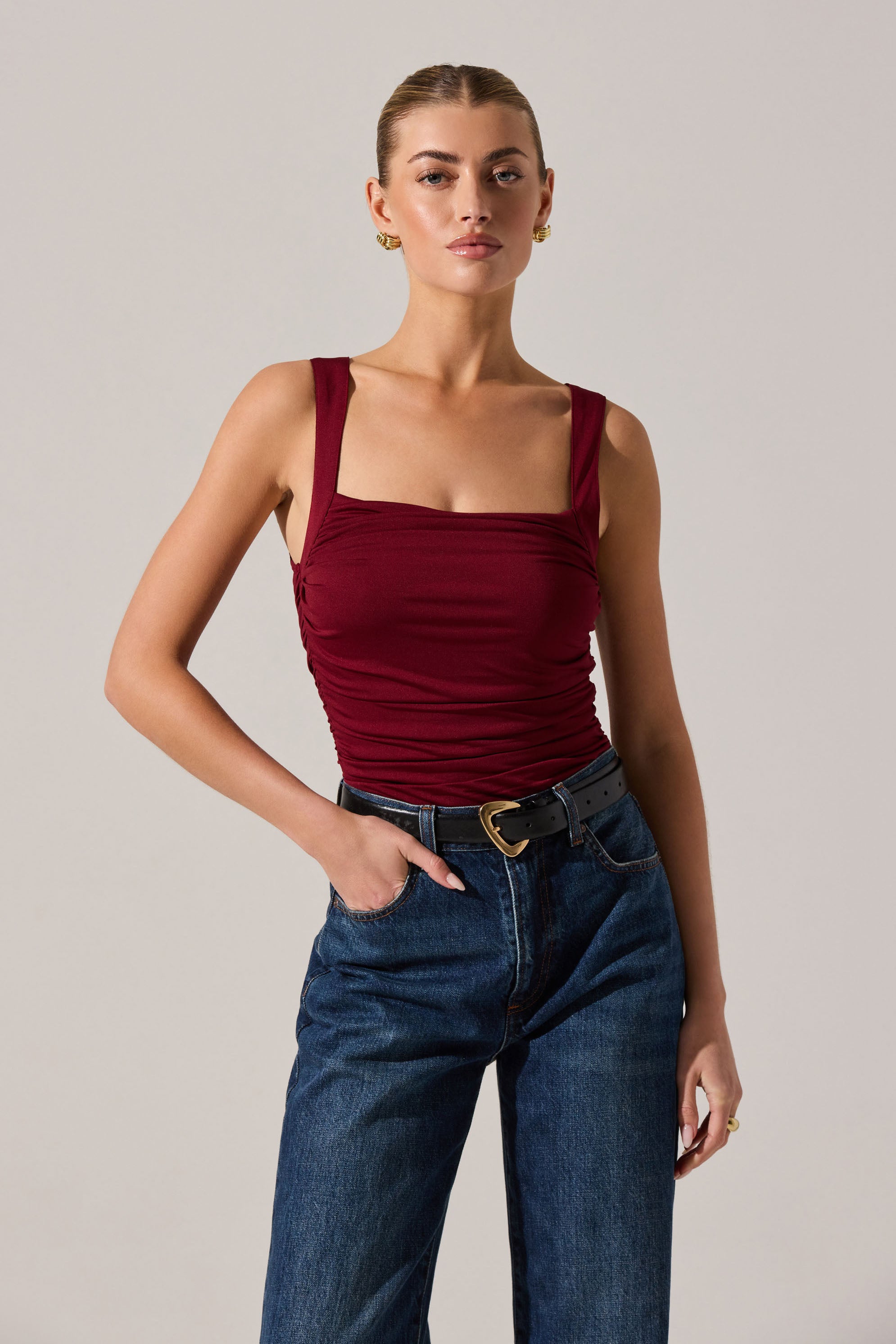 A young woman is wearing a sleeveless, fitted, burgundy top with wide straps and a ruched design across the front. She pairs it with high-waisted, dark-wash jeans and a black belt with a gold heart-shaped buckle.