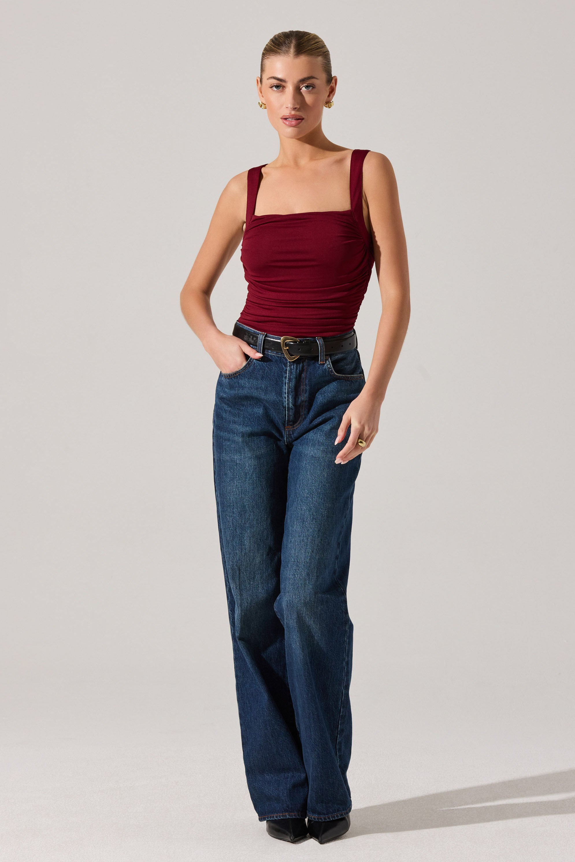 A woman is wearing a fitted, sleeveless, burgundy top with wide shoulder straps and a gathered, ruched design across the chest. She has paired it with high-waisted, dark blue, straight-leg jeans and a black belt with a gold buckle.