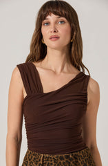 A woman with light skin and shoulder-length brown hair wearing a sleeveless, dark brown, ruched top with a one-shoulder design. She is also wearing gold hoop earrings and a leopard print skirt.