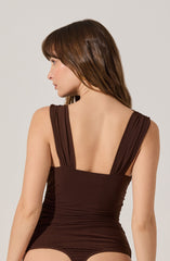 A woman is wearing a sleeveless, dark brown fitted dress with wide, ruched straps that have a gathered appearance at the shoulders. The dress is snug around the waist, creating a smooth, streamlined silhouette.