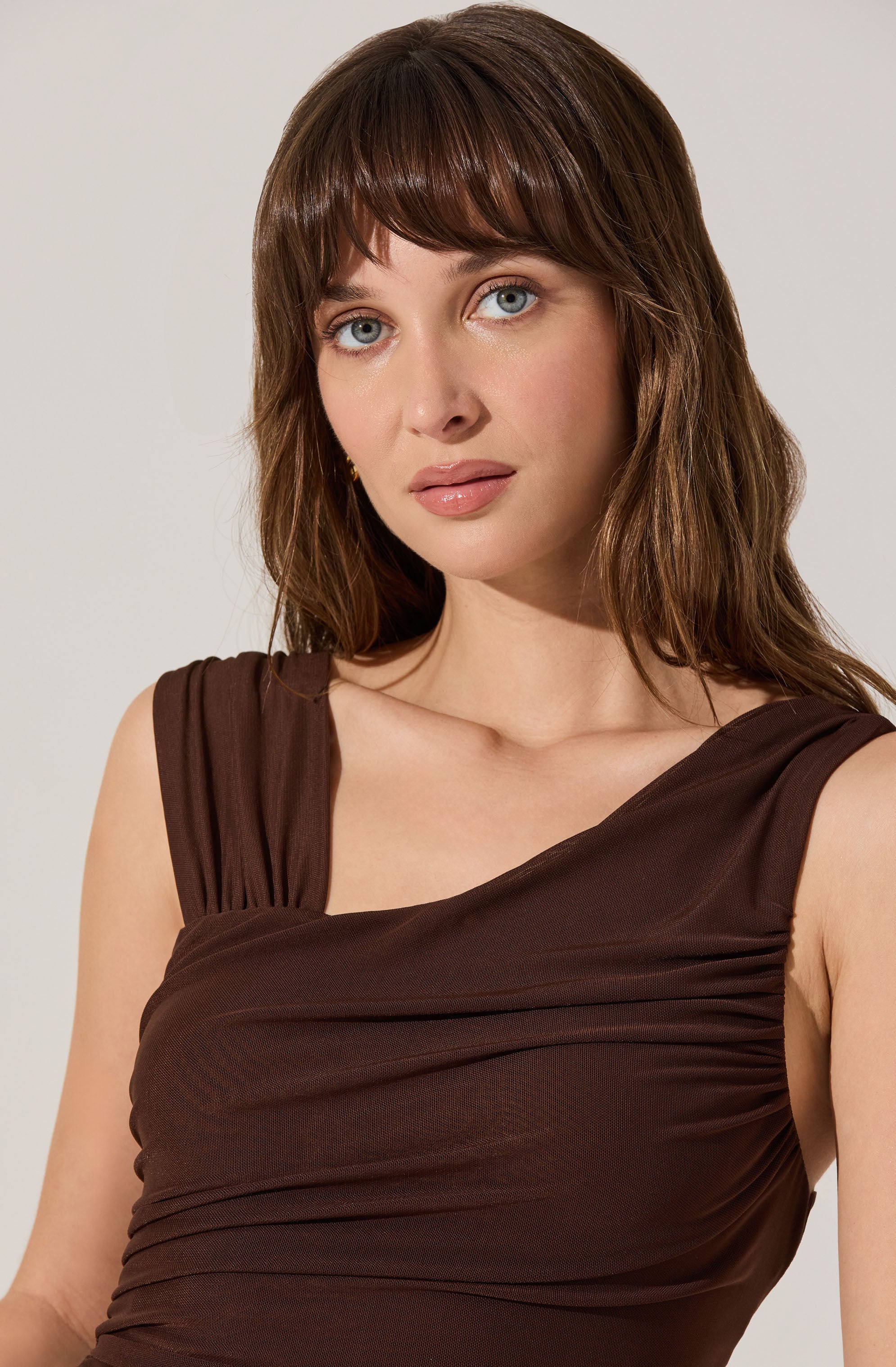 A woman is wearing a sleeveless, dark brown dress with gathered fabric detailing across the bust and shoulders. The dress has wide straps that drape over her shoulders.
