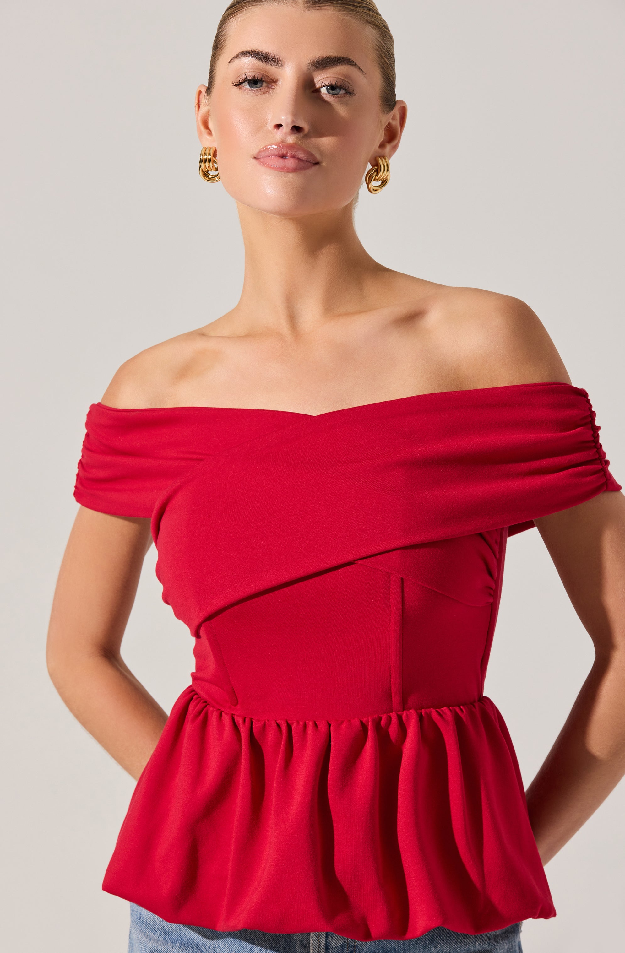 A woman is wearing an off-the-shoulder dress in a bright red color. The dress features a fitted bodice with horizontal ruching along the neckline and a gathered peplum-style waist. She is accessorized with large gold hoop earrings.