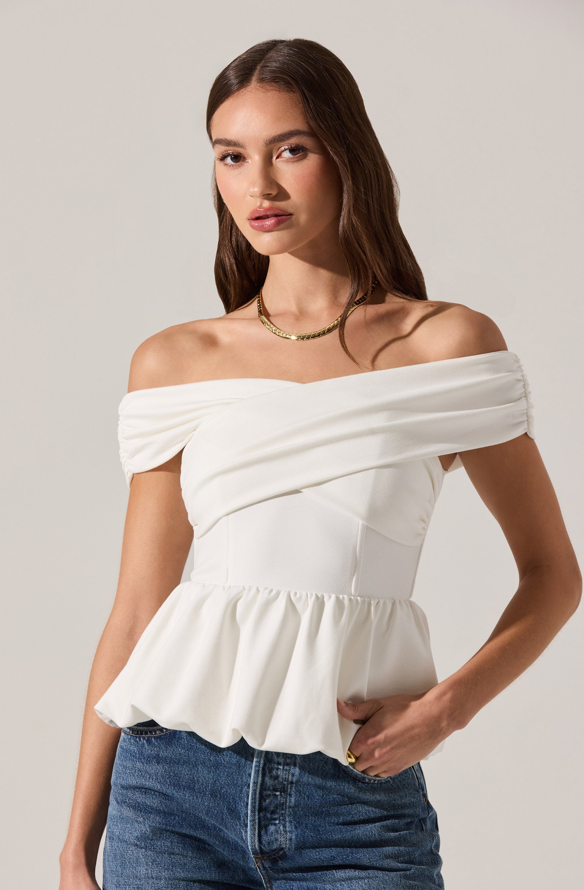 A woman is wearing an off-the-shoulder white top with a draped bodice and a ruffled, gathered hem. The top features wide, ruched sleeves that sit off the shoulders. She pairs it with blue jeans.