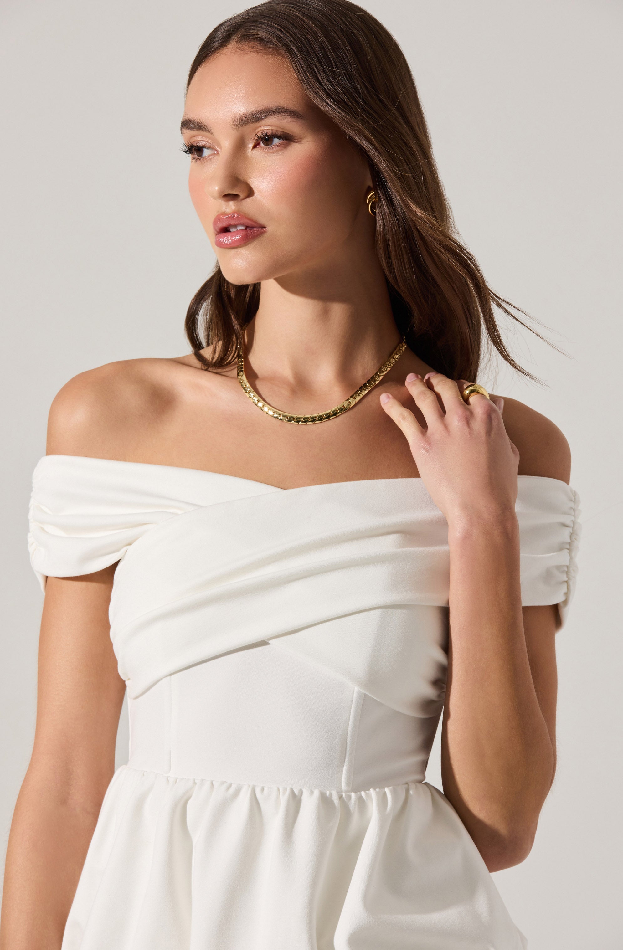 A woman is wearing an off-the-shoulder white dress with ruched fabric at the neckline and a fitted bodice. She has accessorized with a gold necklace, matching earrings, and rings on her fingers. Her hair is styled in loose waves.