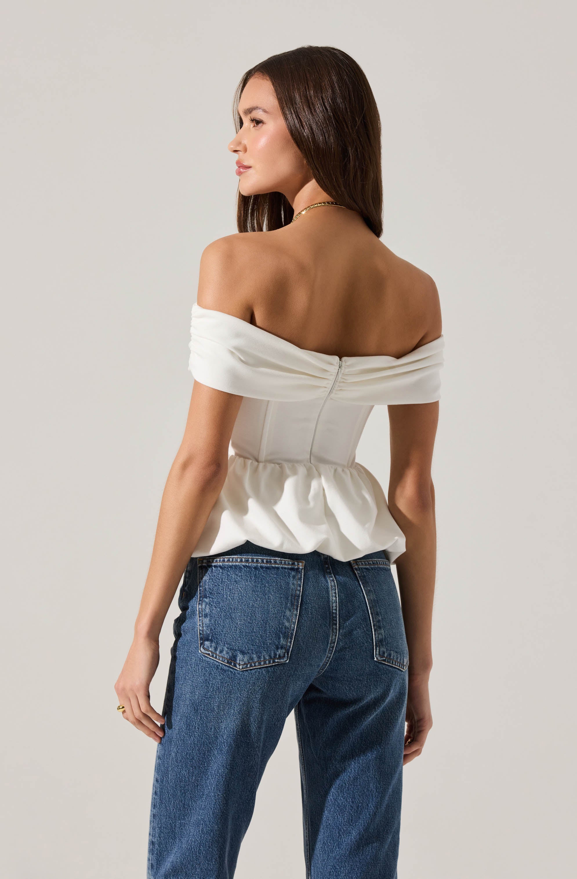 A woman wearing an off-the-shoulder, cream-colored top with a ruched, peplum waist and a back zipper, paired with high-waisted blue jeans.